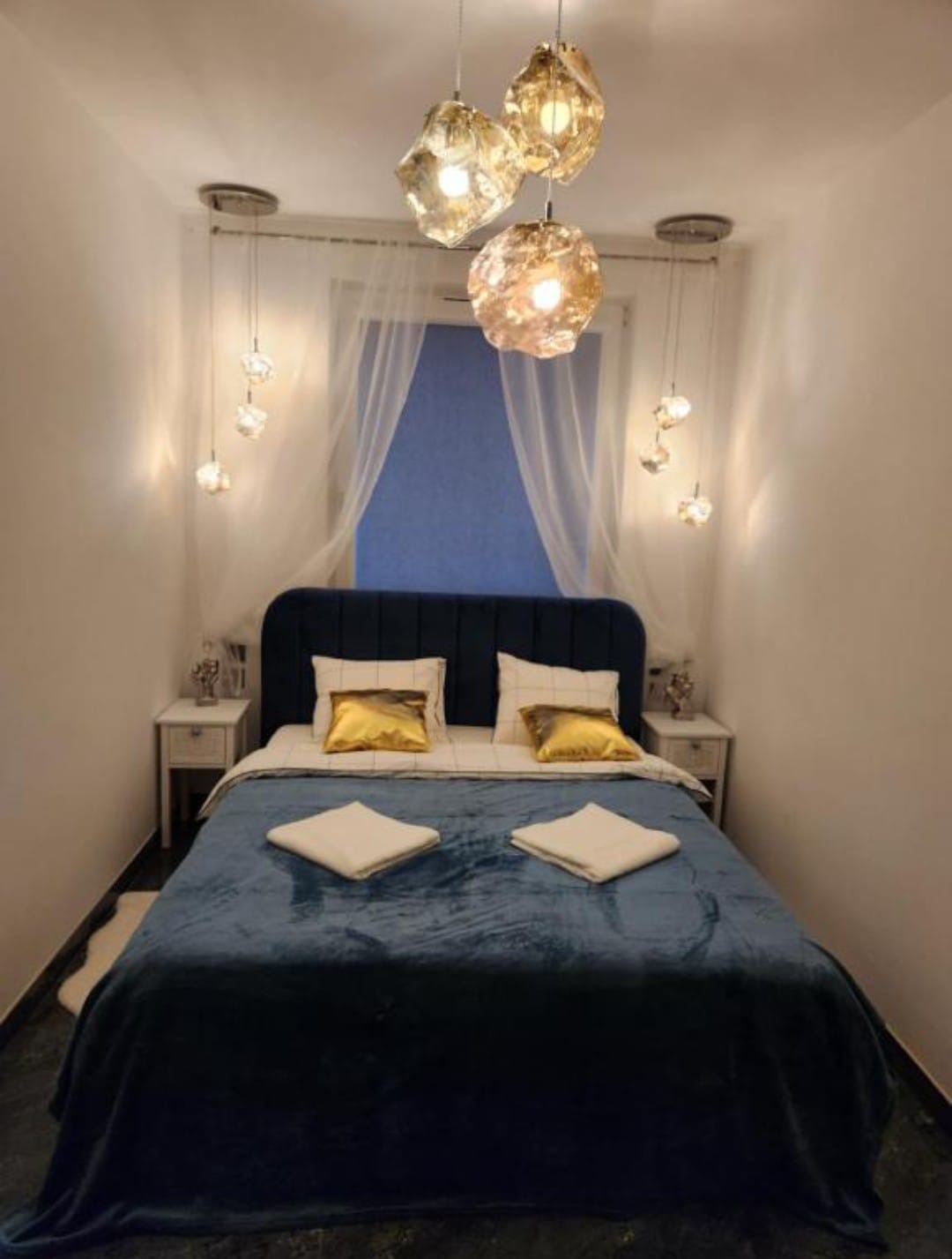 Klimatyczna sypialnia w Gdańsku po wykończeniu: niebieskie łóżko z weluru, złote poduszki, designerskie lampy wiszące i subtelne zasłony w oknie.