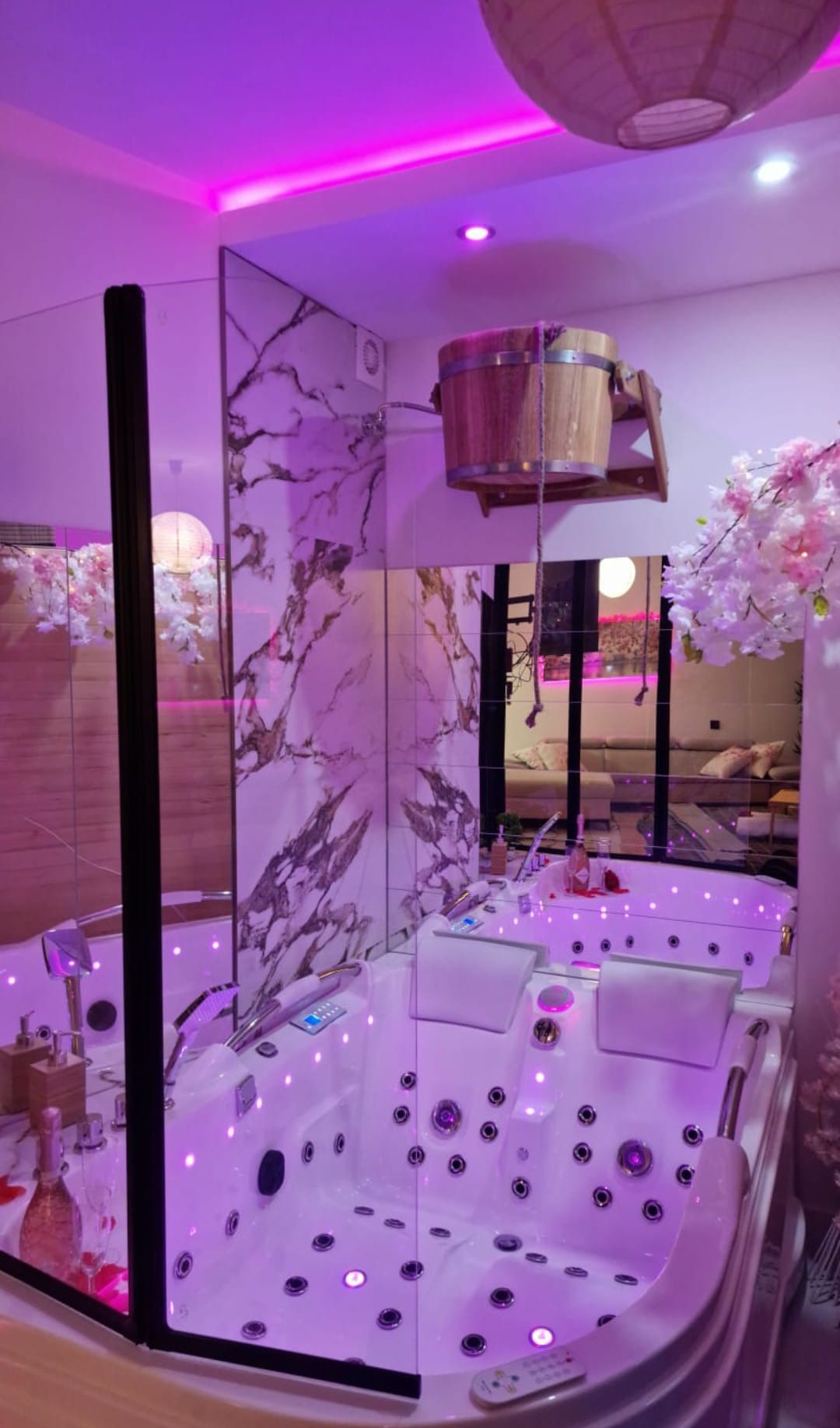 Nowoczesna łazienka z jacuzzi, marmurowe płytki, szklana kabina i dekoracyjny drewniany kubeł. Całość oświetlona fioletowym światłem.