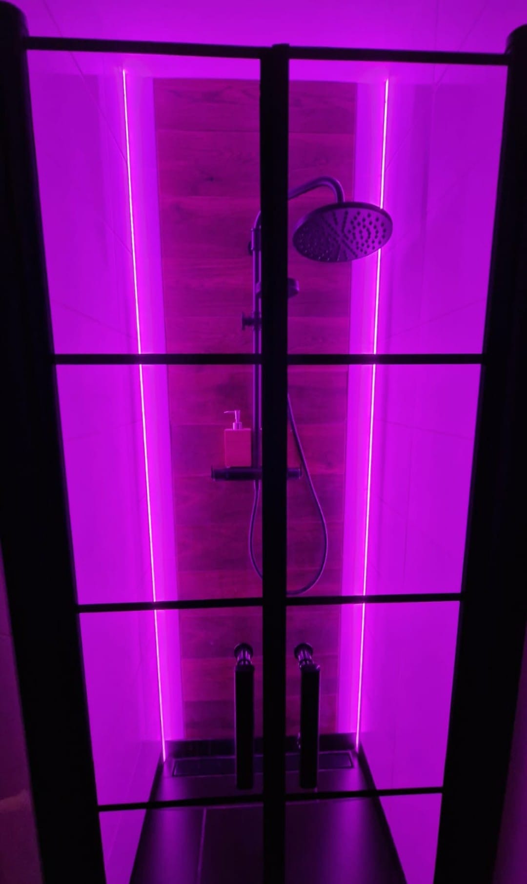Nowoczesna kabina prysznicowa z deszczownicą, podświetlona fioletowym światłem LED, z elementami drewnopodobnymi na ścianie i czarnymi, metalowymi detalami.