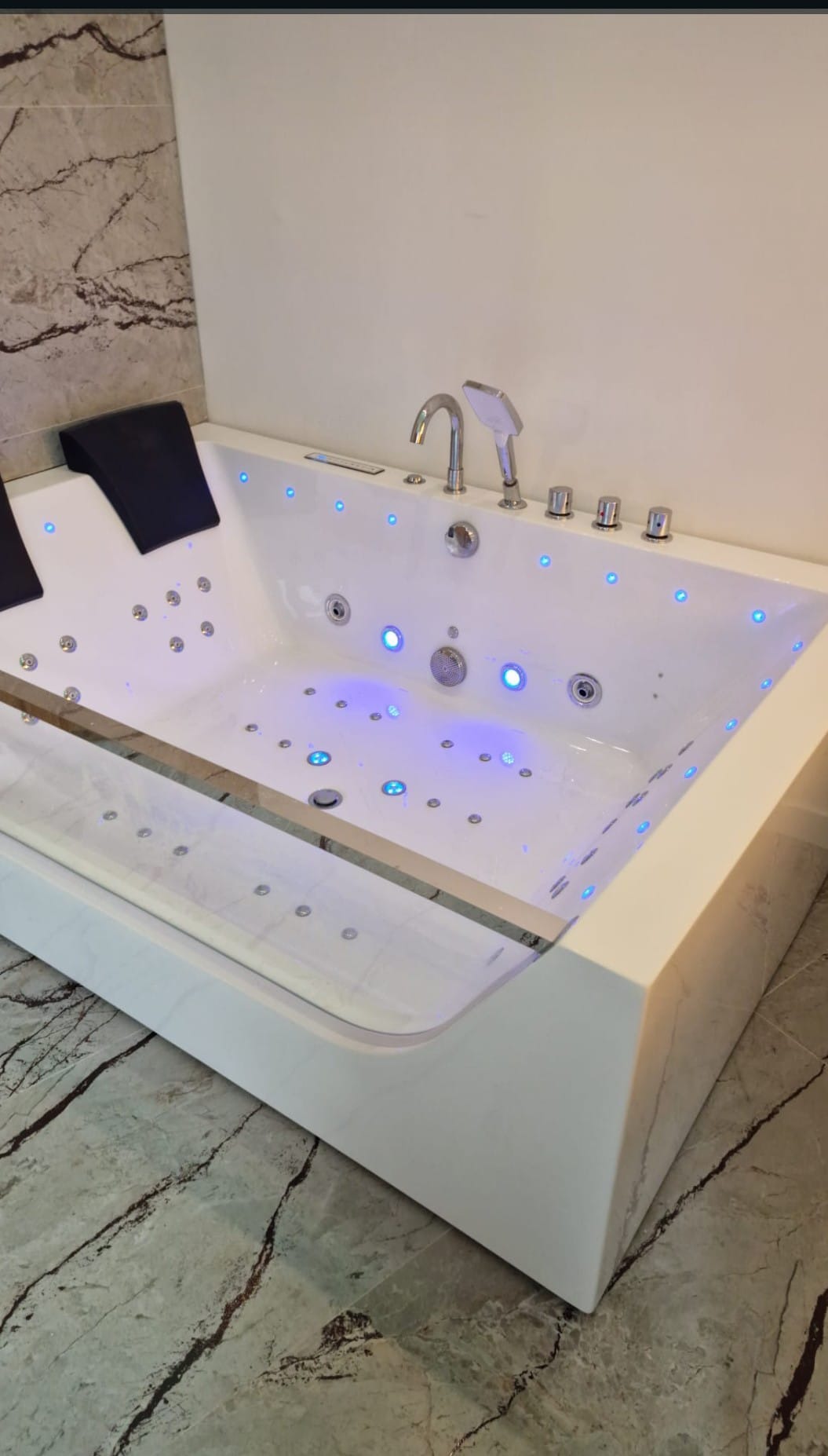 Nowoczesna, biała wanna z hydromasażem i podświetleniem LED, z czarnymi zagłówkami, zamontowana w łazience z marmurową ścianą i podłogą. Widok z góry.