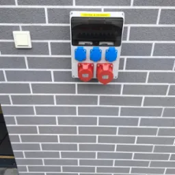 Zewnętrzna skrzynka elektryczna z gniazdami siłowymi (czerwone i niebieskie) zamontowana na szarej ścianie imitującej cegłę. Widoczny też włącznik światła.