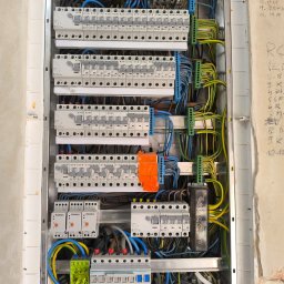 MElektryka - Rozdzielnica elektryczna w trakcie montażu, widoczne liczne przewody, bezpieczniki i listwy zaciskowe. Elementy marki Eaton i Kanlux. Ściana z notatkami.