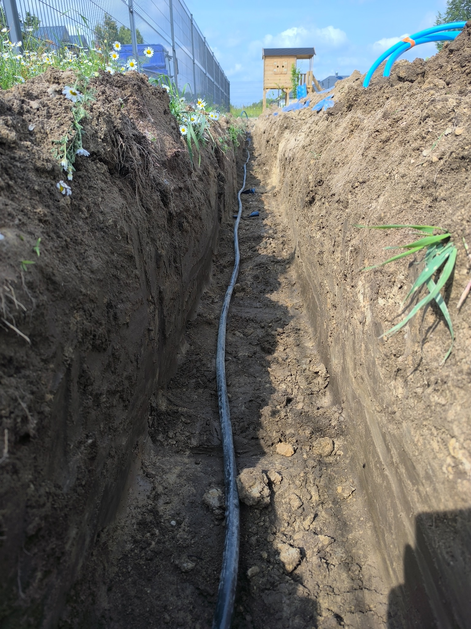 Wkopana w ziemię rura instalacji elektrycznej, widoczna w wykopanym rowie. W tle ogrodzenie i plac zabaw.
