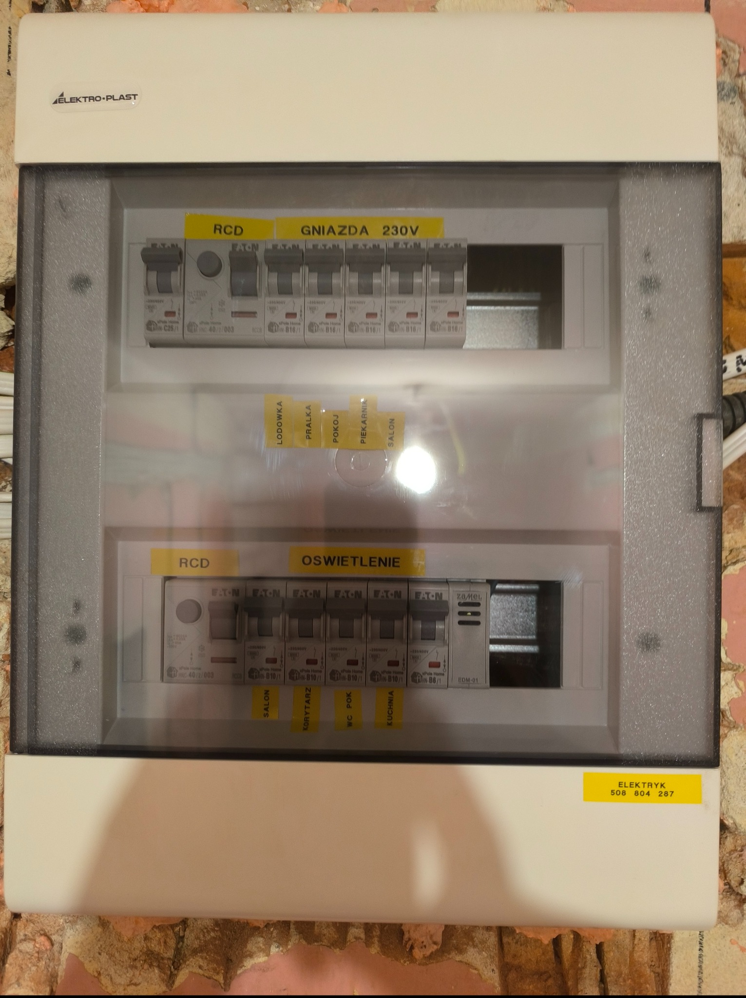 Rozdzielnia elektryczna z bezpiecznikami i oznaczeniami obwodów (gniazda, oświetlenie, lodówka, pralka, pokój). Tabliczka z napisem 'Elektryk' i numerem telefonu.