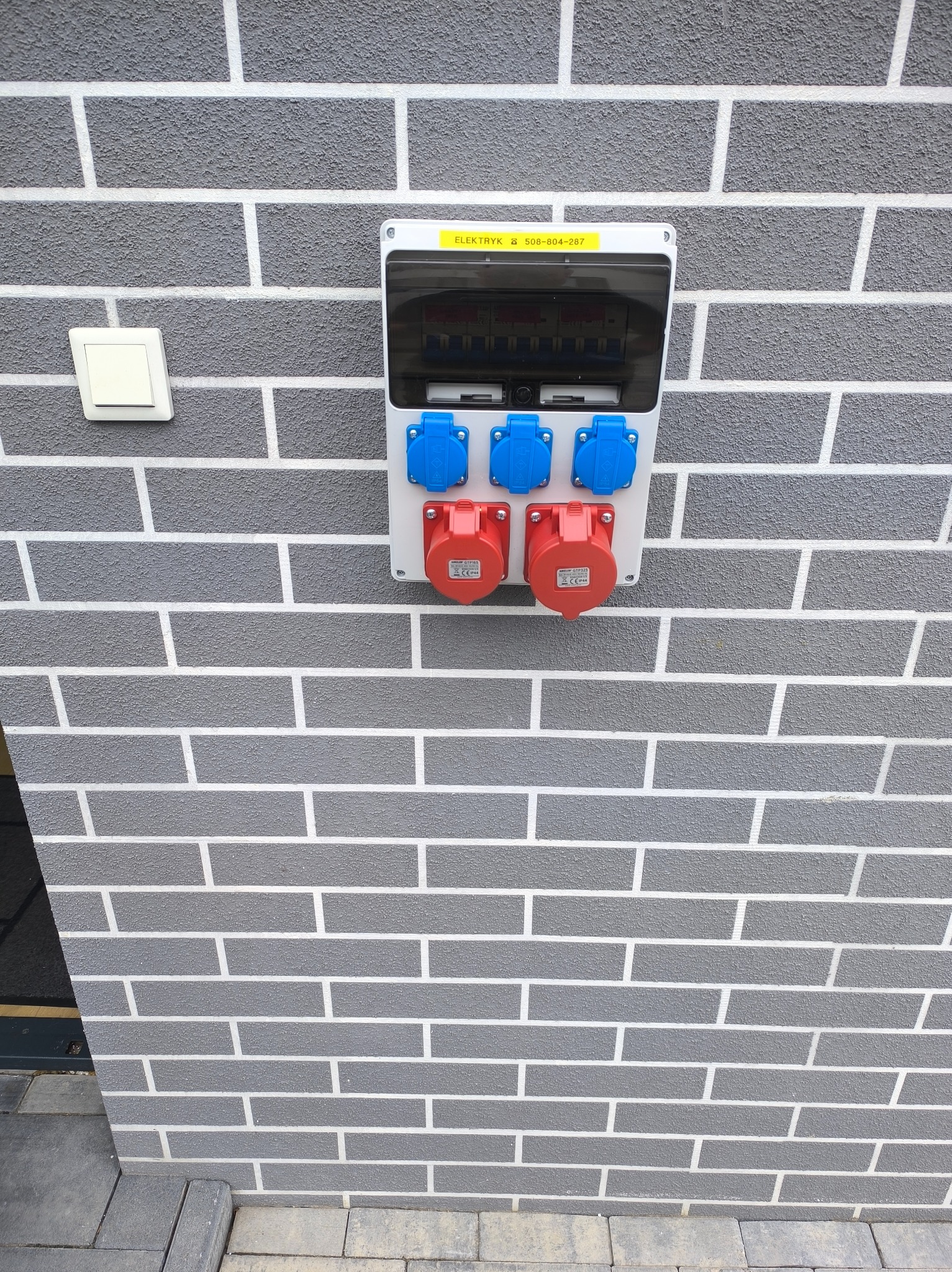 Zewnętrzna skrzynka elektryczna z gniazdami siłowymi (czerwone i niebieskie) zamontowana na szarej ścianie imitującej cegłę. Widoczny też włącznik światła.