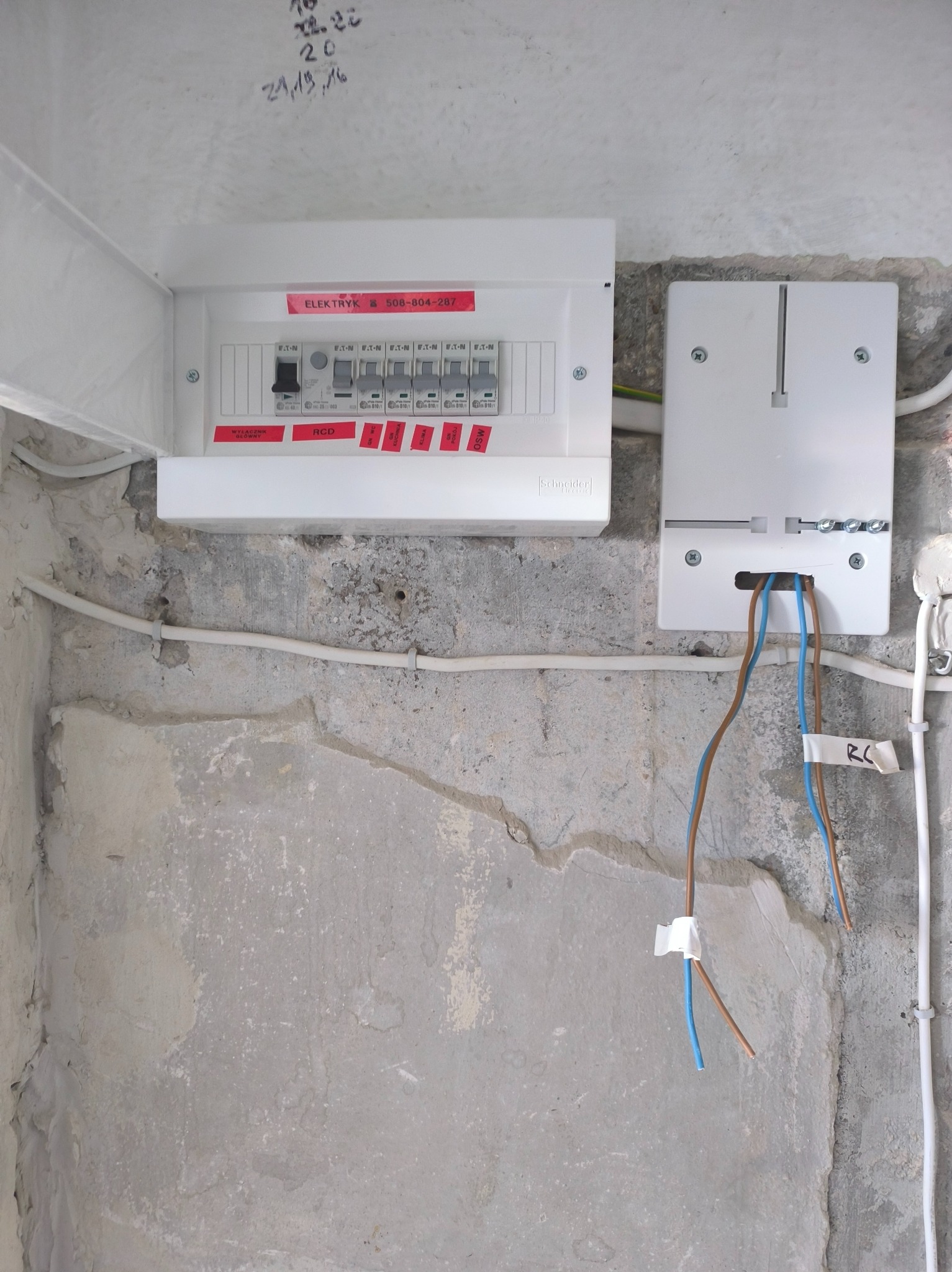 Otwarta skrzynka elektryczna z bezpiecznikami i naklejką z numerem telefonu elektryka, obok druga puszka z wystającymi kablami na tle surowej ściany.