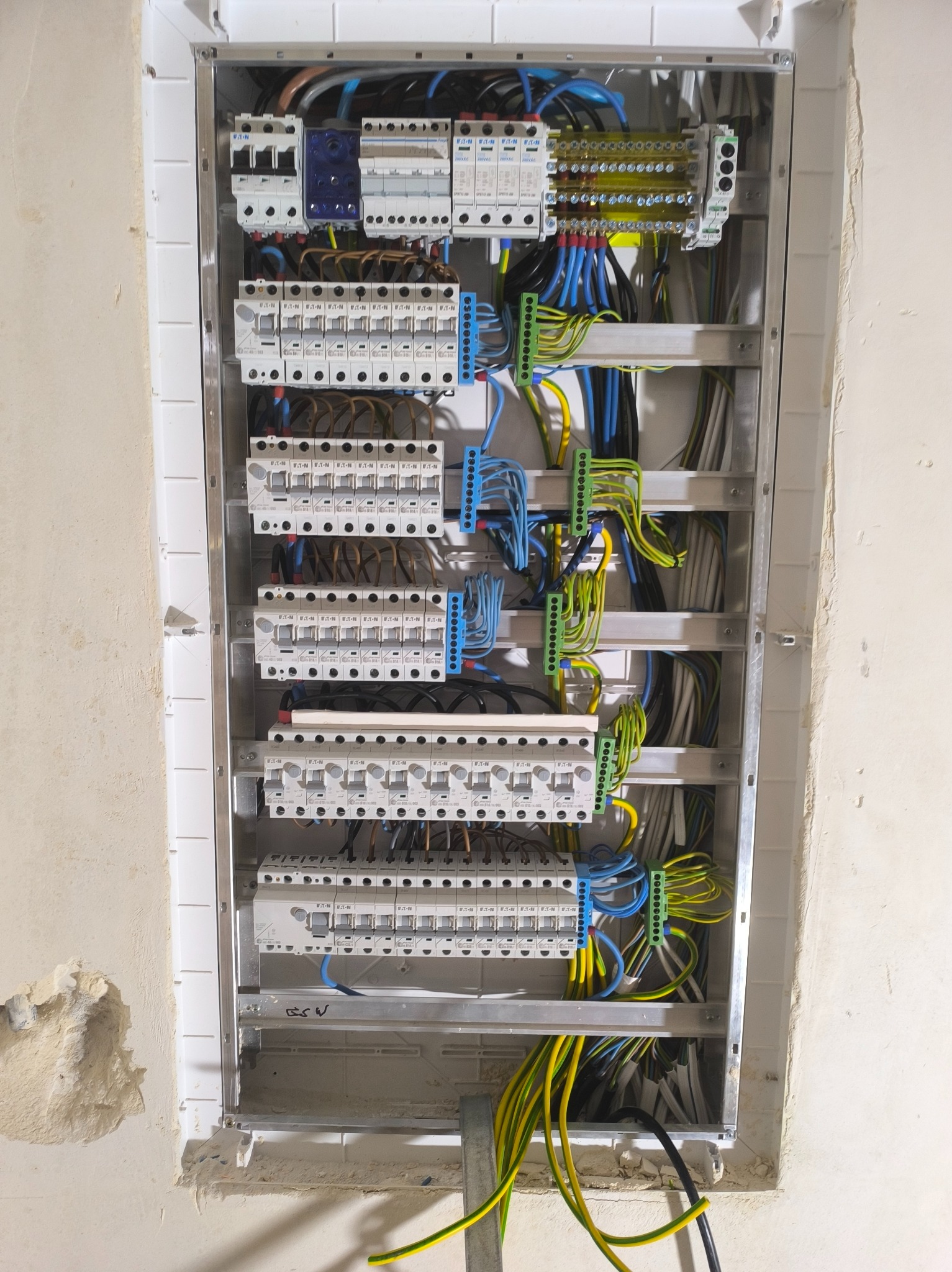 Otwarta skrzynka elektryczna z zainstalowanymi bezpiecznikami i kolorowymi przewodami na ścianie w surowym stanie, widoczne uszkodzenia tynku.