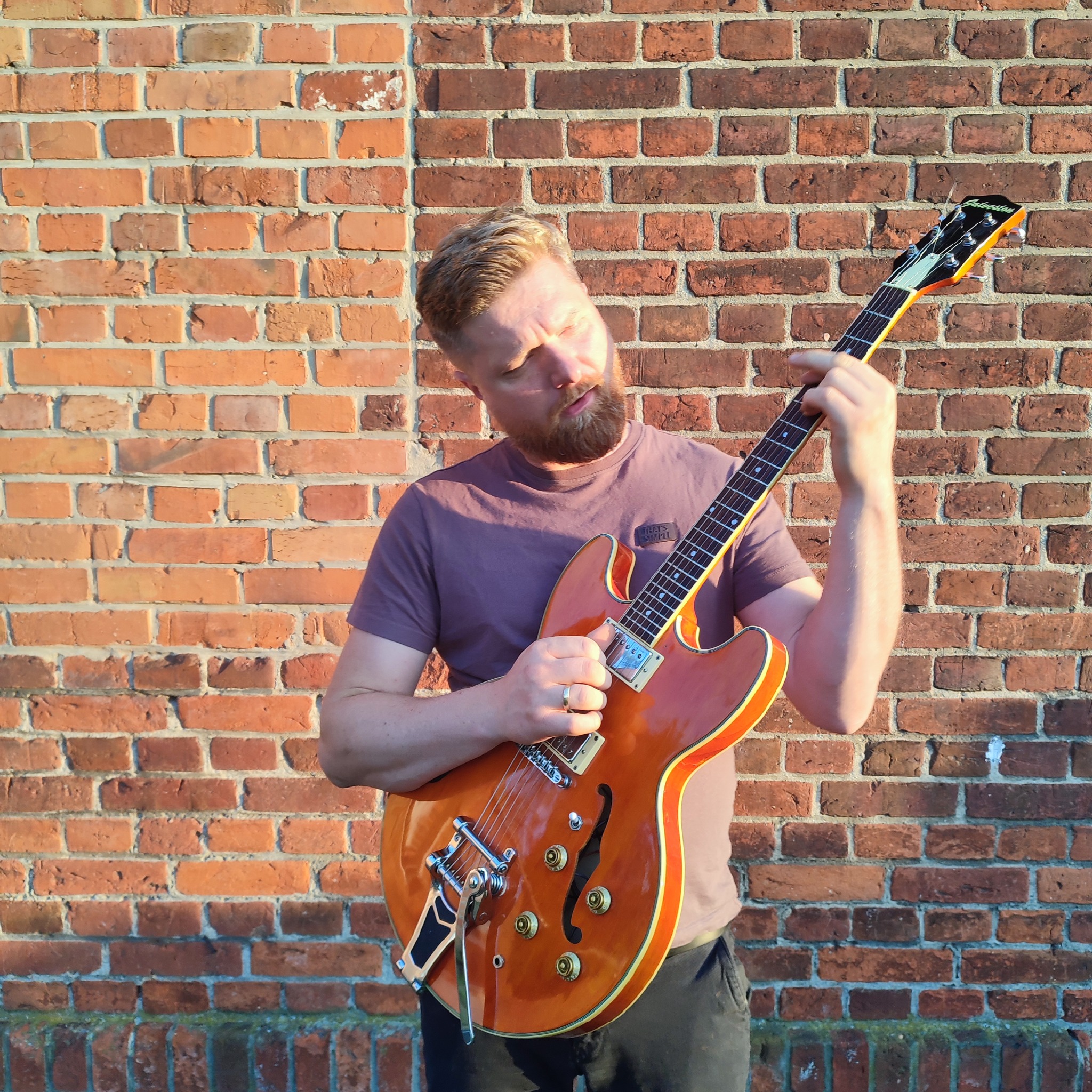 Mężczyzna z brodą gra na pomarańczowej gitarze elektrycznej na tle ceglanej ściany w słoneczny dzień. Artysta skupiony na grze.