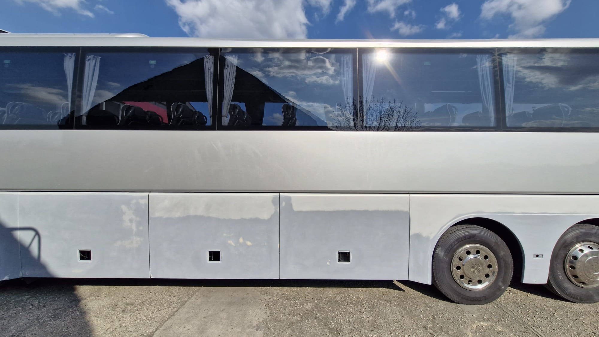 Autobus w trakcie naprawy blacharskiej. Widoczne szpachlowane panele boczne, przygotowane do lakierowania. Szyby odbijają błękitne niebo z chmurami.