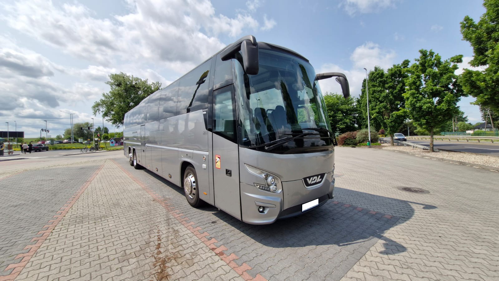 Srebrny autokar VDL na parkingu z kostki brukowej, widok z przodu, błękitne niebo z chmurami w tle. Pojazd po serwisie blacharsko-lakierniczym.