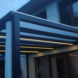 ADMT Serwis Adrian Filipczuk - Nowoczesna pergola z oświetleniem LED, zamontowana przy domu. Widoczne elementy konstrukcyjne i ciepłe światło lamp. Zdjęcie z dołu, wieczorową porą.