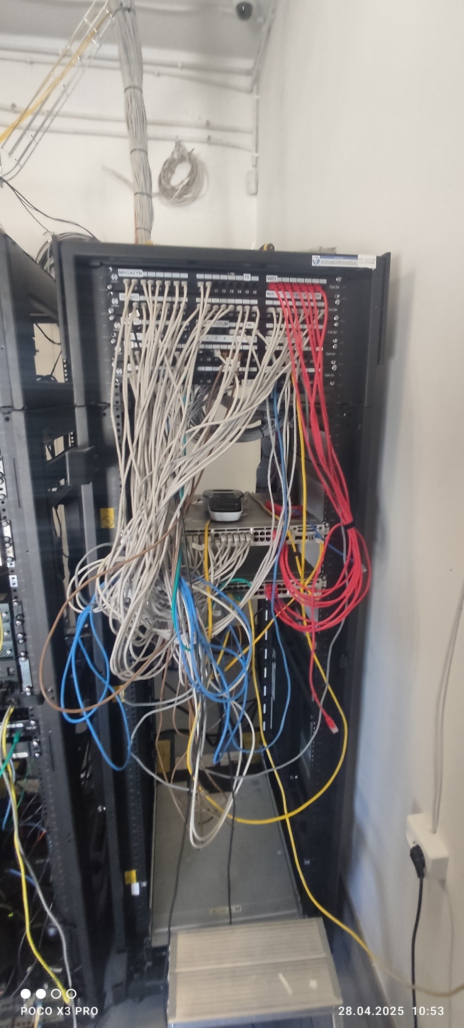 Szafa rack z widocznym panelem krosowniczym i chaotycznym okablowaniem sieciowym (kable Ethernet w różnych kolorach) oraz elementami aktywnej sieci.