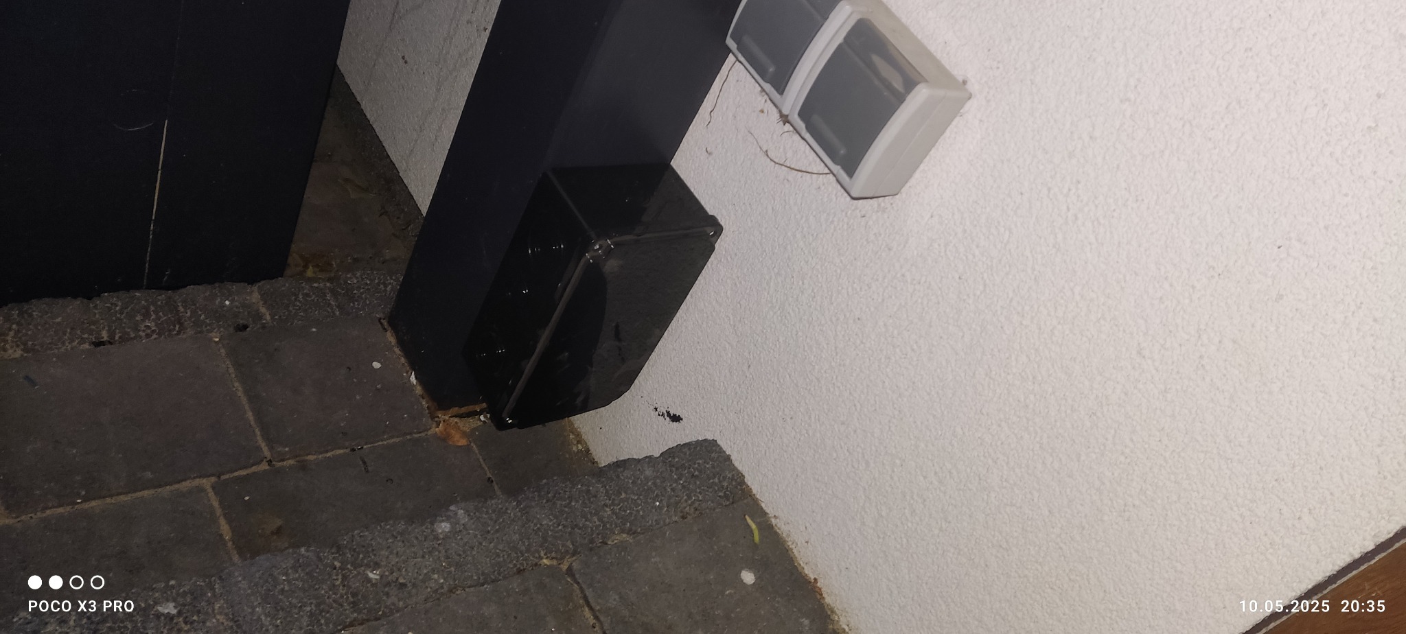 Zewnętrzna lampa LED na białej ścianie obok czarnego słupka, na tle szarej kostki brukowej. Nocne oświetlenie elewacji budynku.