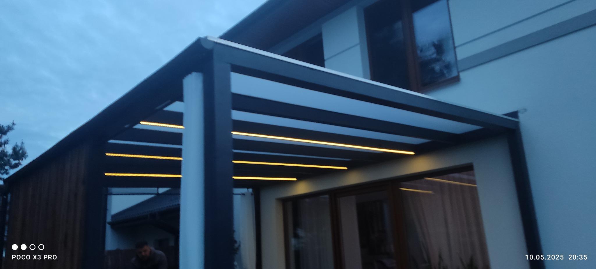 Nowoczesna pergola z oświetleniem LED, zamontowana przy domu. Widoczne elementy konstrukcyjne i ciepłe światło lamp. Zdjęcie z dołu, wieczorową porą.