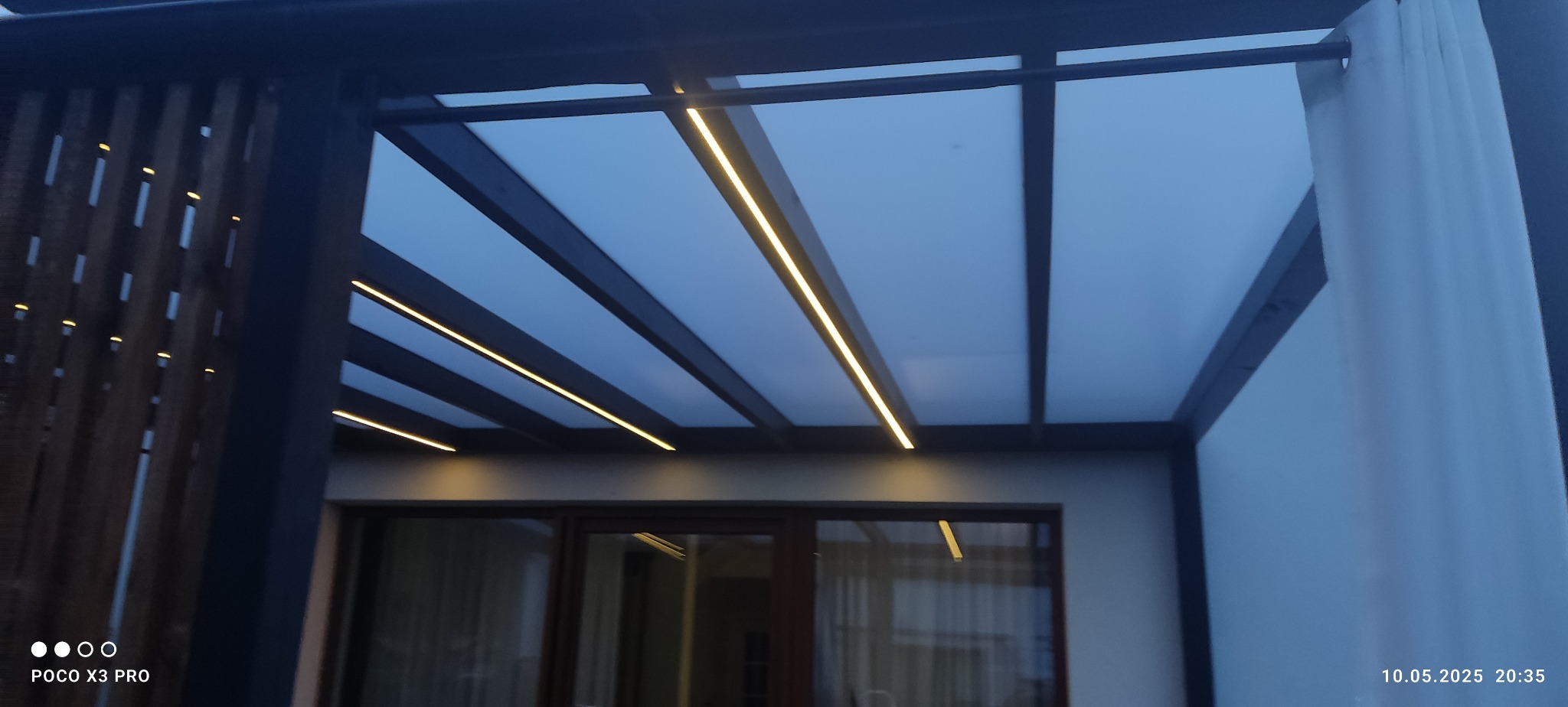 Zadaszenie tarasu z oświetleniem LED, widok od dołu. Nowoczesna konstrukcja z drewnianymi elementami i zasłoną. Elegancki design.