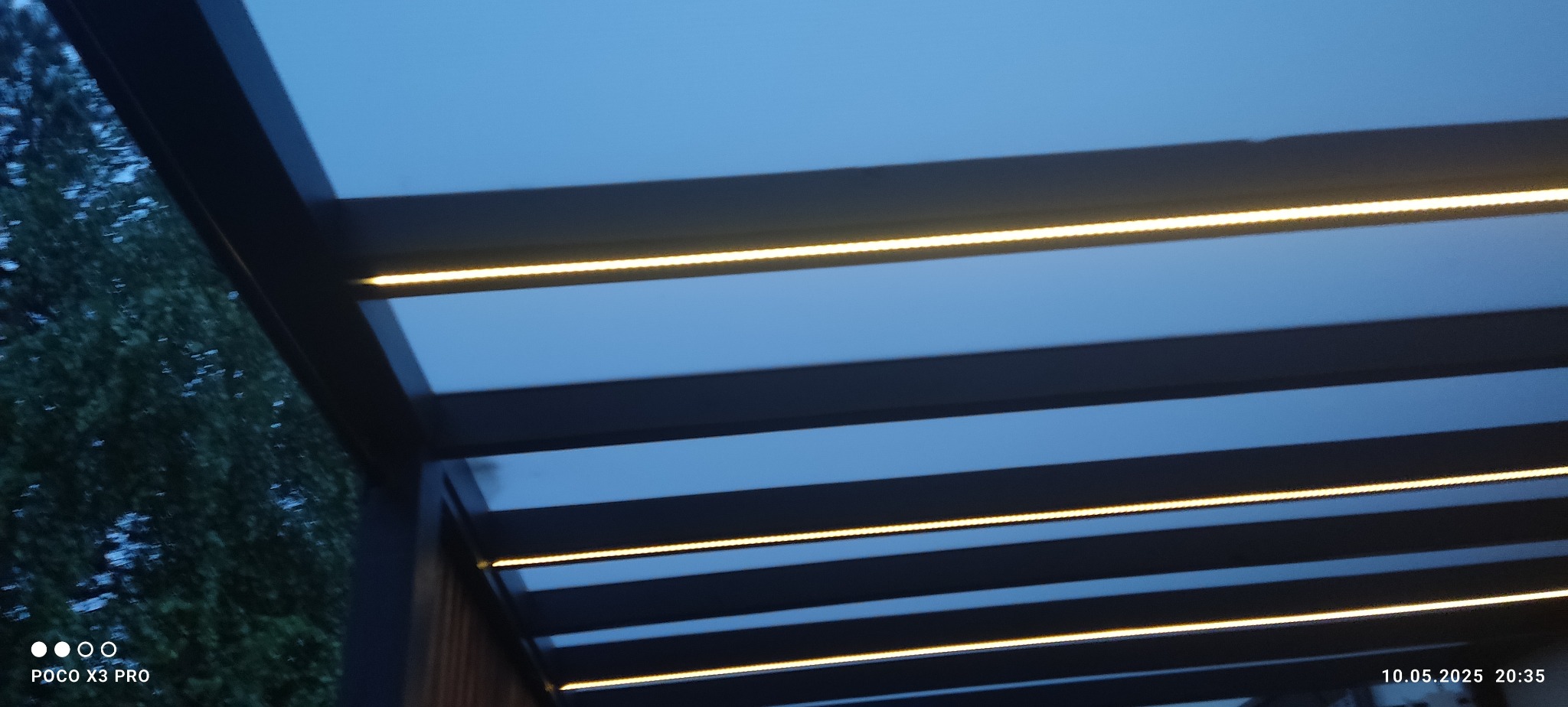 Nowoczesna pergola z oświetleniem LED wbudowanym w konstrukcję. Widok z dołu, oświetlenie tworzy ciepłe linie na tle nieba i ciemnych belek.