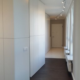 JAKUB SZELĄG PROJEKTOWANIE WNĘTRZ - Biały przedpokój z zabudową szaf wnękowych pod sufit, oświetleniem szynowym i ciemną podłogą. Widok na białe drzwi wejściowe i okna z grzejnikami.