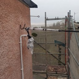 JAKUB JASIŃSKI - Zabezpieczone elementy instalacji odgromowej na ścianie budynku, widoczne rusztowanie i uchwyt uziemiający. Przewody owinięte folią i taśmą izolacyjną.