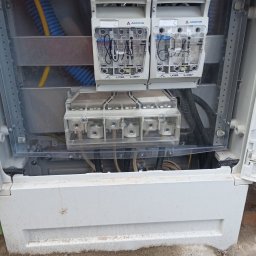 JAKUB JASIŃSKI - Otwarta skrzynka elektryczna z bezpiecznikami i złączami. Widoczne kable i przewody. Elementy instalacji elektrycznej marki Apator. Rozłączniki RBK00 i złącza szynowe.