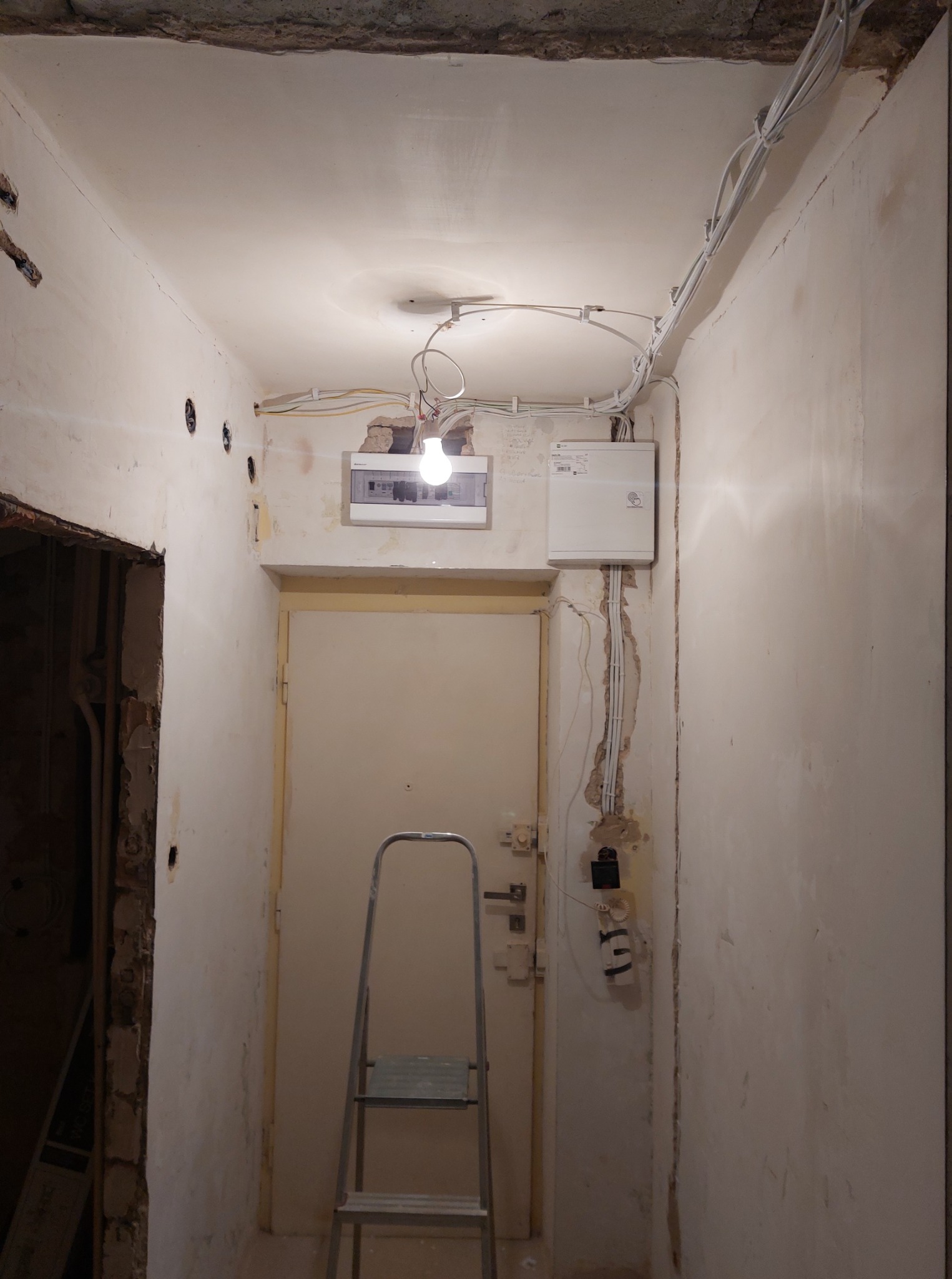 Korytarz w trakcie remontu: widoczne kable elektryczne na suficie, skrzynka elektryczna na ścianie, drabina, ściany w stanie surowym.