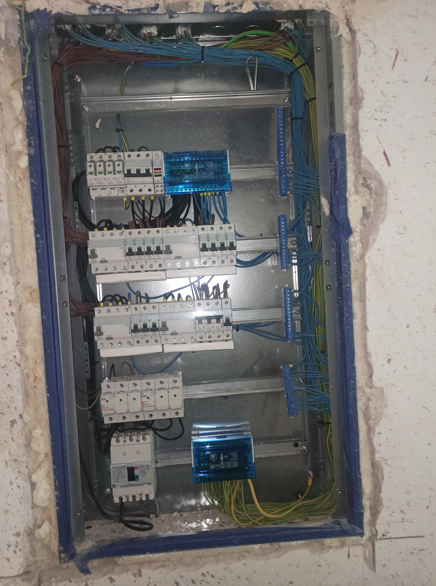 Otwarta, metalowa skrzynka z bezpiecznikami i przewodami w kolorach: brązowym, niebieskim i zielono-żółtym, zamontowana w ścianie z widocznymi śladami po kuciu.