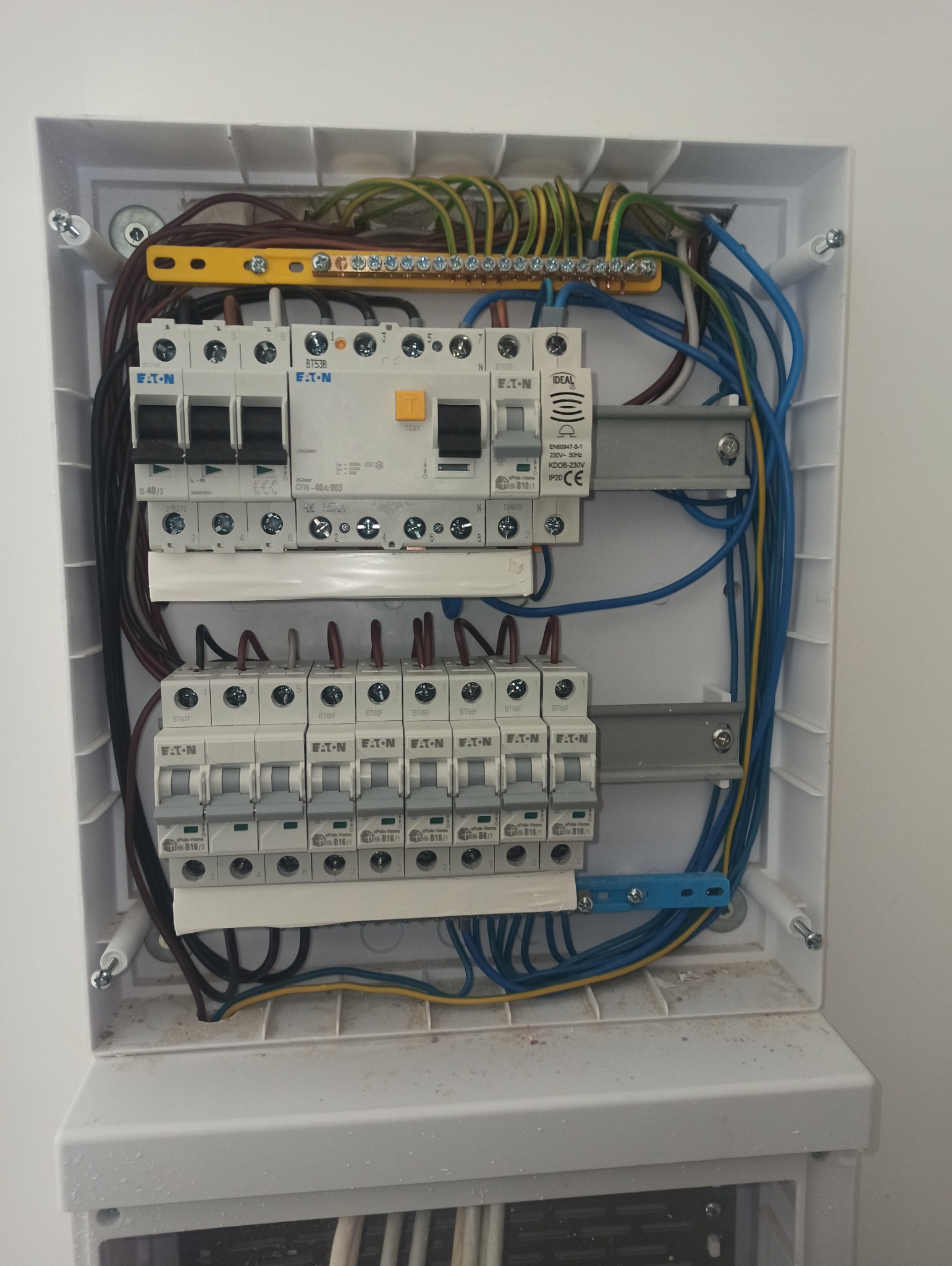 Rozdzielnica elektryczna w białej obudowie z widocznymi zabezpieczeniami, przewodami i elementami montażowymi. Widok z bliska na wnętrze skrzynki.