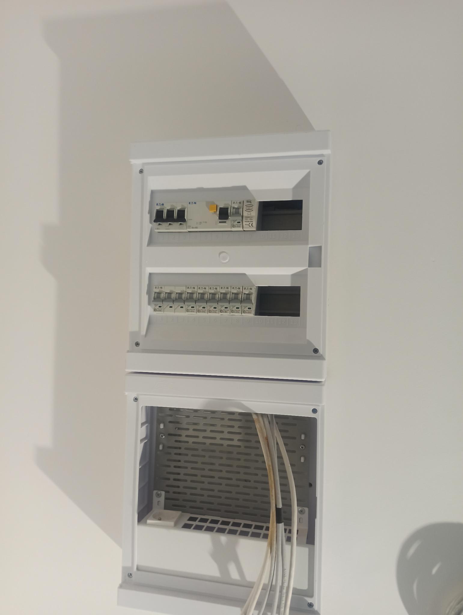 Biała, dwuczęściowa skrzynka rozdzielcza Eaton zamontowana na białej ścianie. Widoczne bezpieczniki i przewody w dolnej części obudowy.