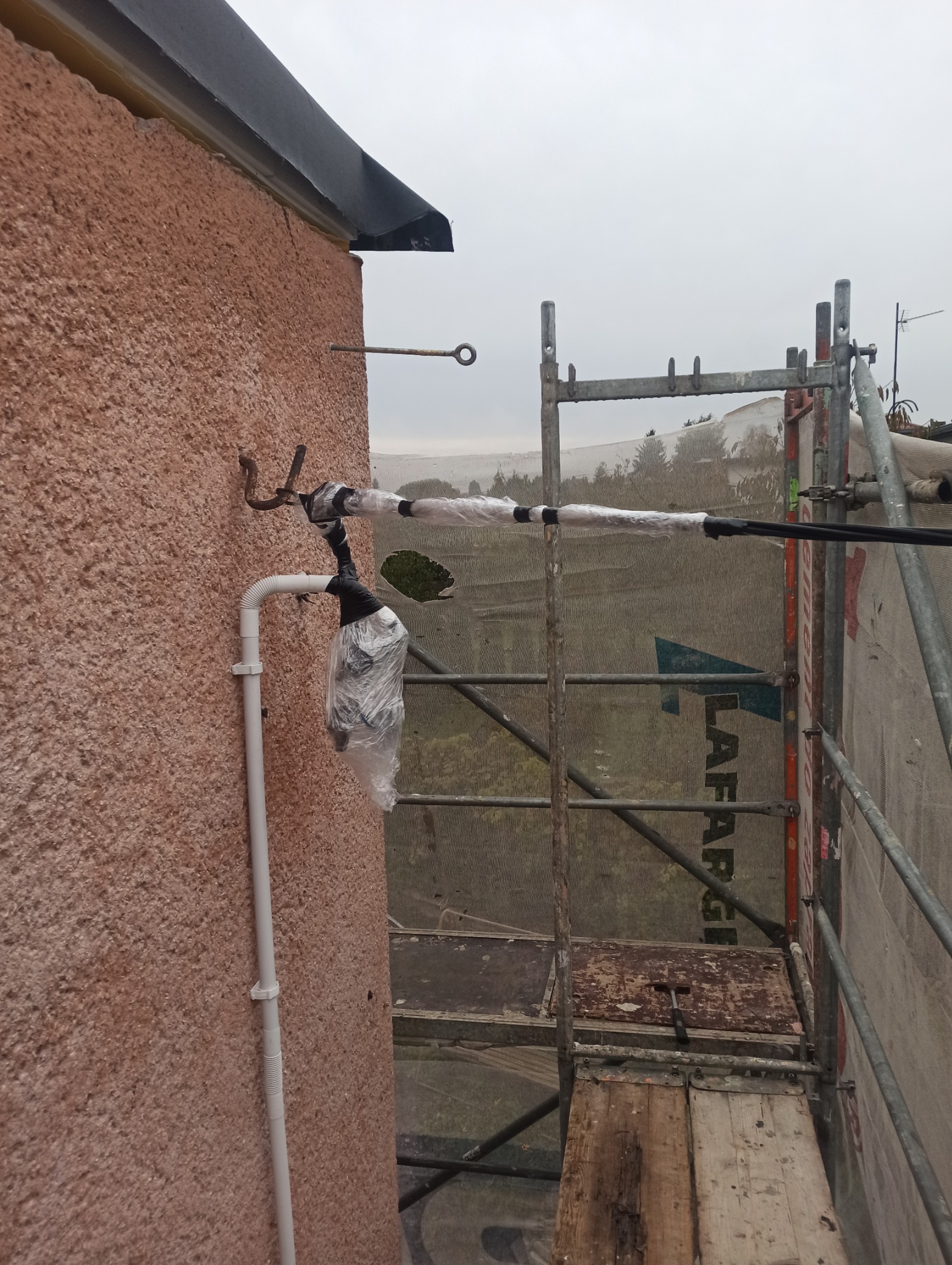 Zabezpieczone elementy instalacji odgromowej na ścianie budynku, widoczne rusztowanie i uchwyt uziemiający. Przewody owinięte folią i taśmą izolacyjną.