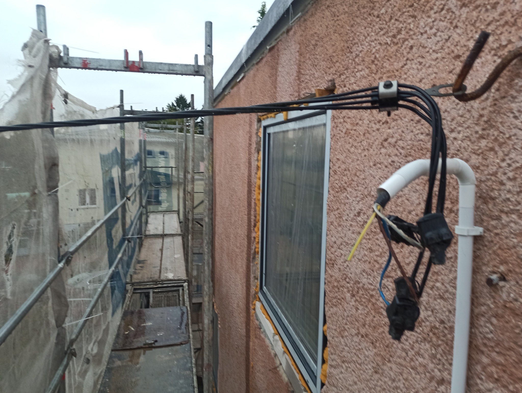 Czarny kabel elektryczny przymocowany do ściany budynku z oknem, widoczne rusztowanie obok. Elementy instalacji elektrycznej na zewnątrz budynku.