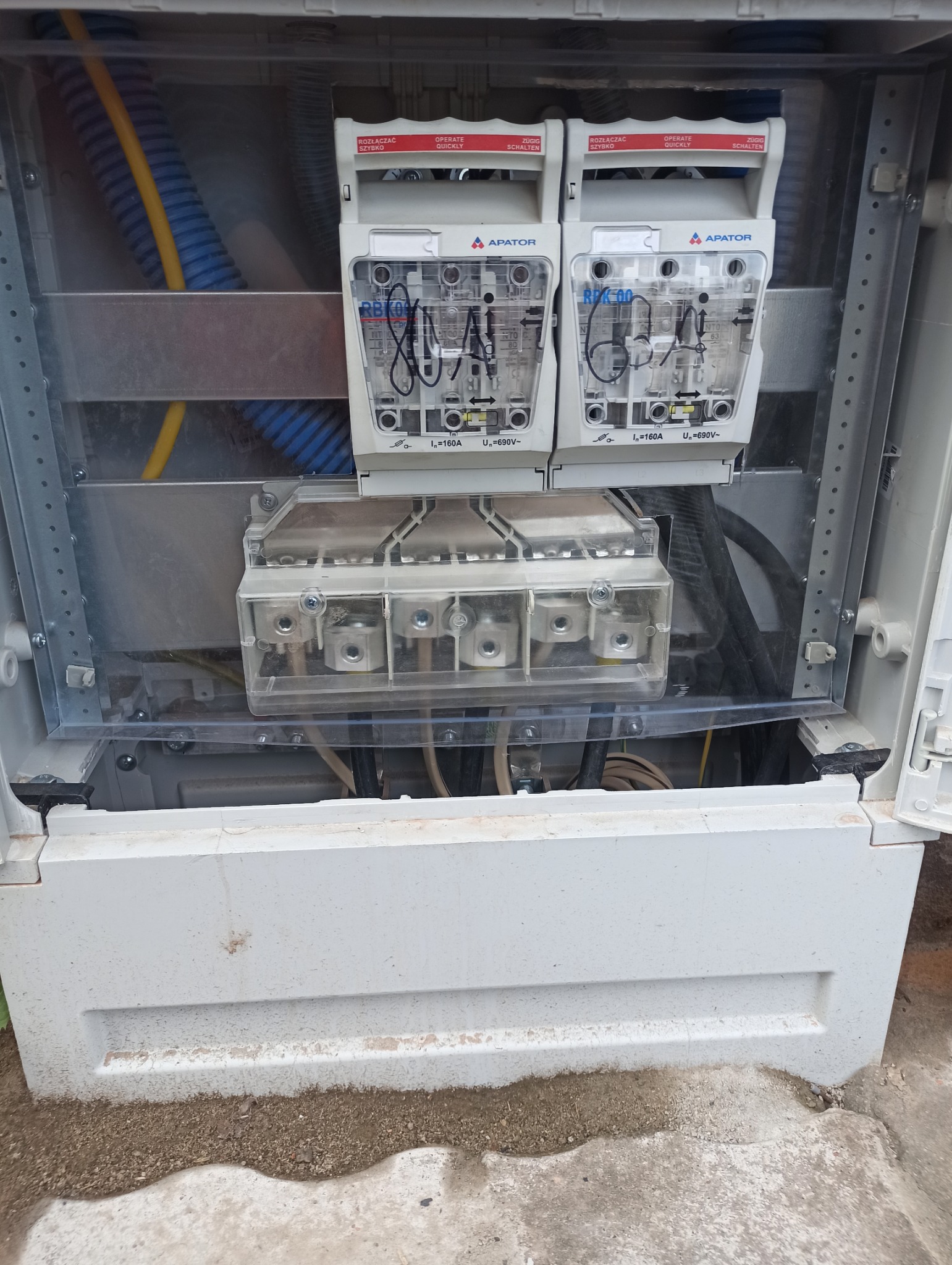 Otwarta skrzynka elektryczna z bezpiecznikami i złączami. Widoczne kable i przewody. Elementy instalacji elektrycznej marki Apator. Rozłączniki RBK00 i złącza szynowe.
