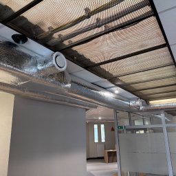 KLIMALA - Instalacja wentylacyjna z aluminiowymi rurami w biurze, widoczne kratki wentylacyjne w suficie podwieszanym i przeszklone ściany działowe.