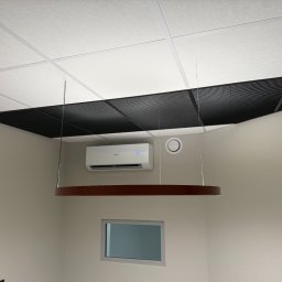 KLIMALA - Biała klimatyzacja Haier zamontowana na ścianie pod sufitem rastrowym w biurze, widoczna okrągła lampa wisząca i małe okno.