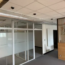 Biuro z widoczną instalacją wentylacyjną pod sufitem, szklane ścianki działowe z matową folią, widoczne czujniki dymu i oświetlenie.