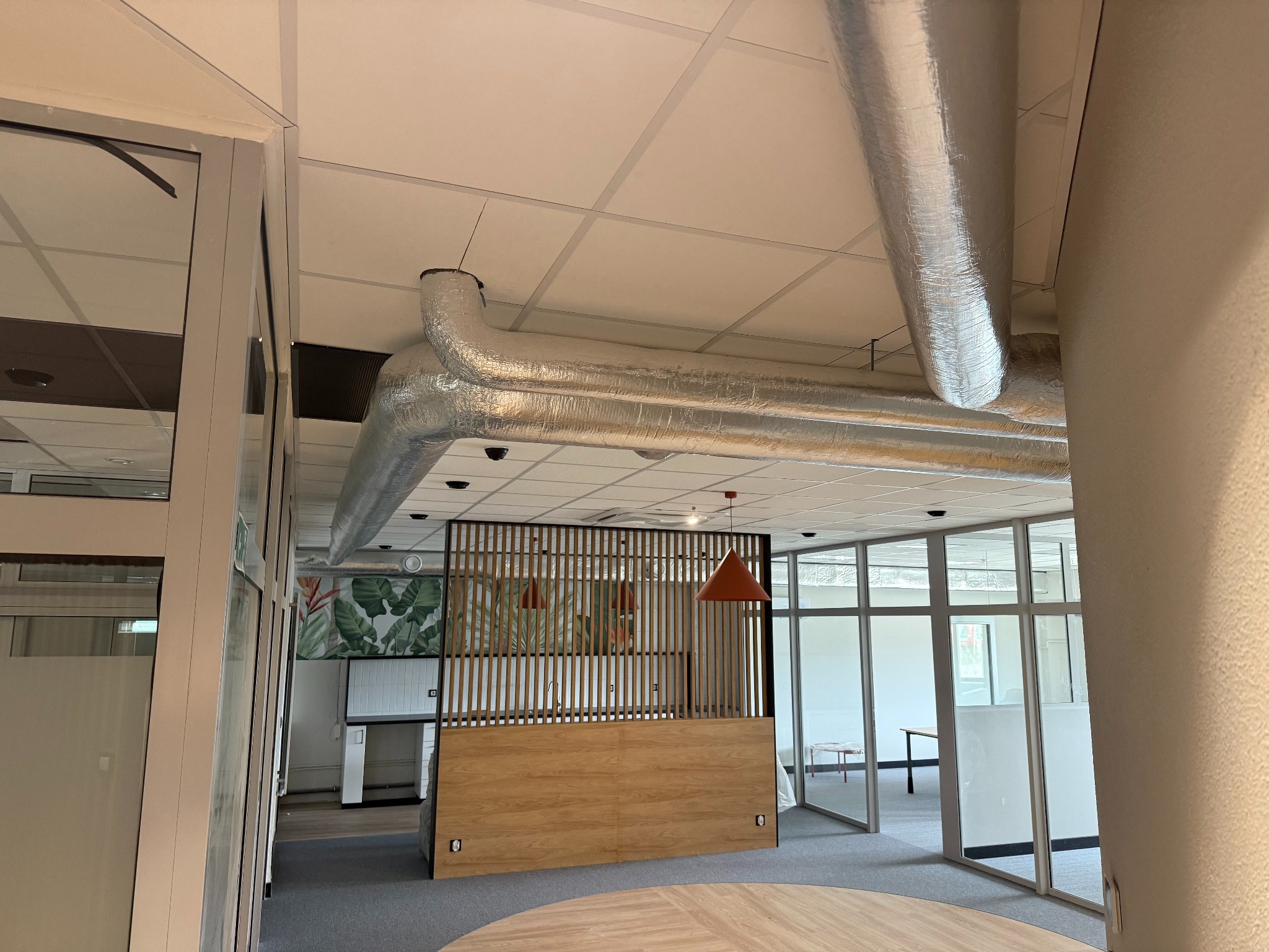 Instalacja wentylacyjna w biurze: srebrne rury pod sufitem, widok z dołu, przeszklone ściany działowe i dekoracyjna ścianka działowa z drewnianymi lamelami.