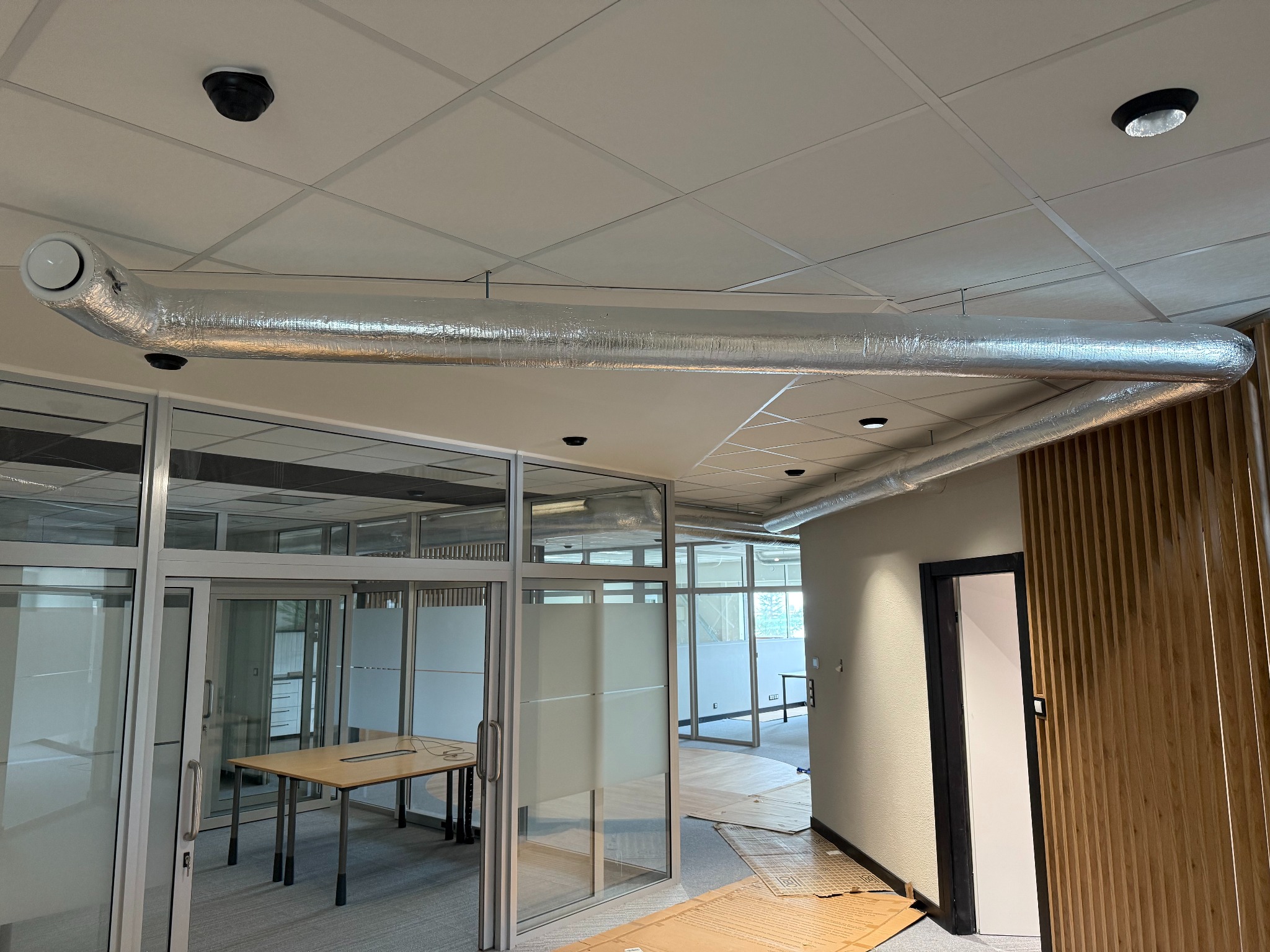 Instalacja wentylacyjna z okrągłym wylotem w biurze, widoczna na suficie podwieszanym, z fragmentami szklanych ścianek działowych i biurek w tle.