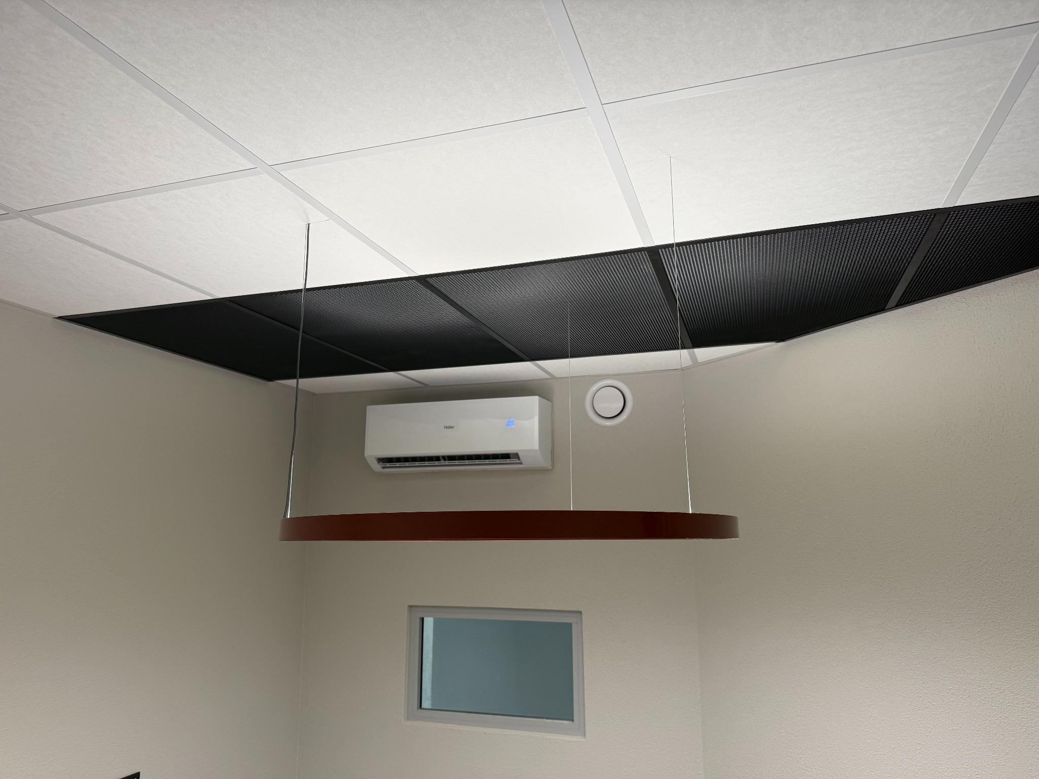 Biała klimatyzacja Haier zamontowana na ścianie pod sufitem rastrowym w biurze, widoczna okrągła lampa wisząca i małe okno.