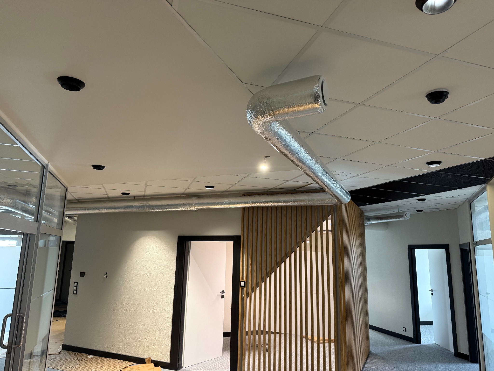 Białe biuro z widoczną instalacją wentylacyjną pod sufitem, oświetleniem punktowym i drewnianą ścianką działową. Nowoczesny design.