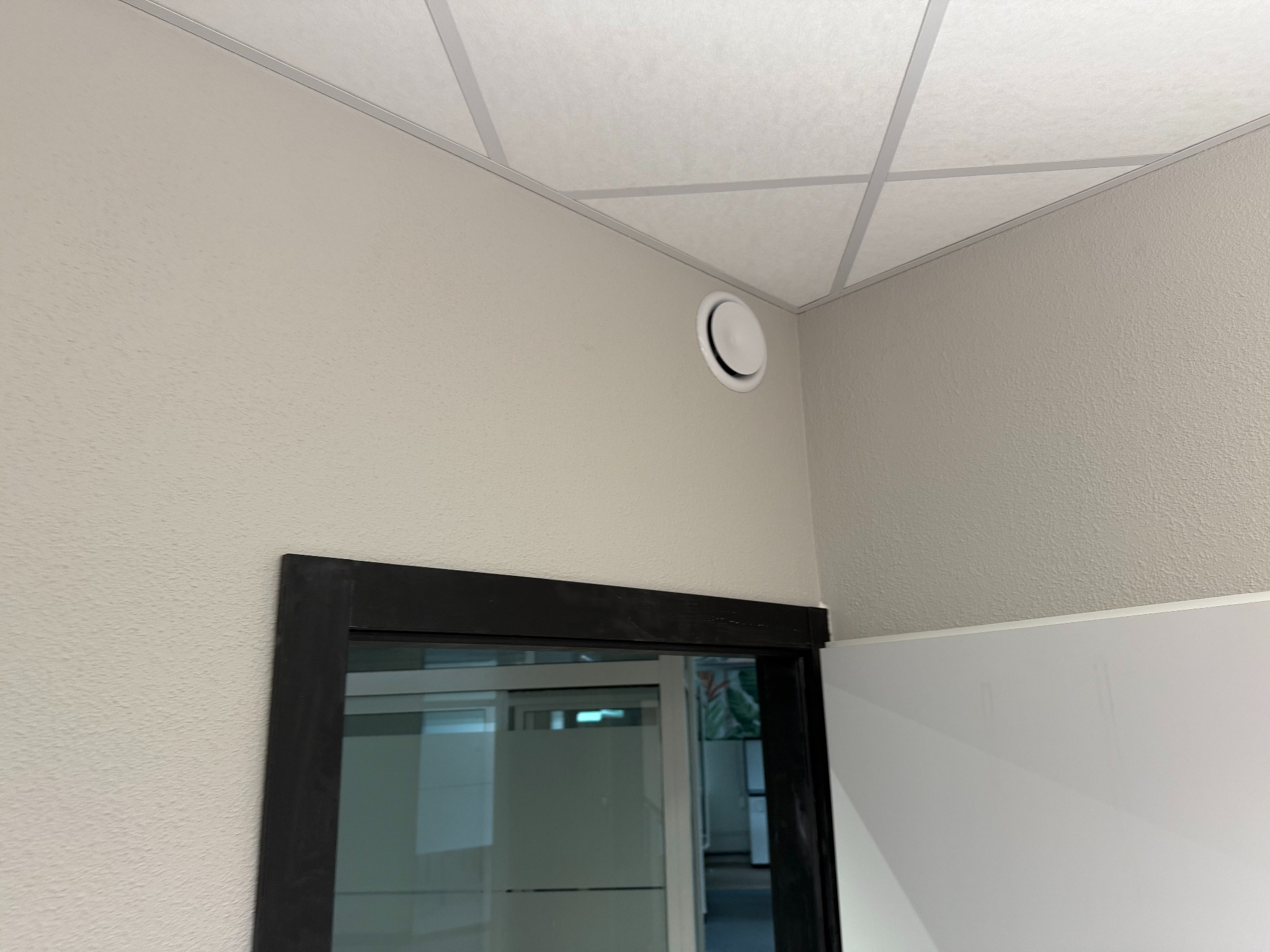 Biały nawiewnik wentylacyjny na ścianie w biurze w Gdyni, widok z dołu, fragment sufitu podwieszanego i ramy drzwiowej. Minimalistyczny design.