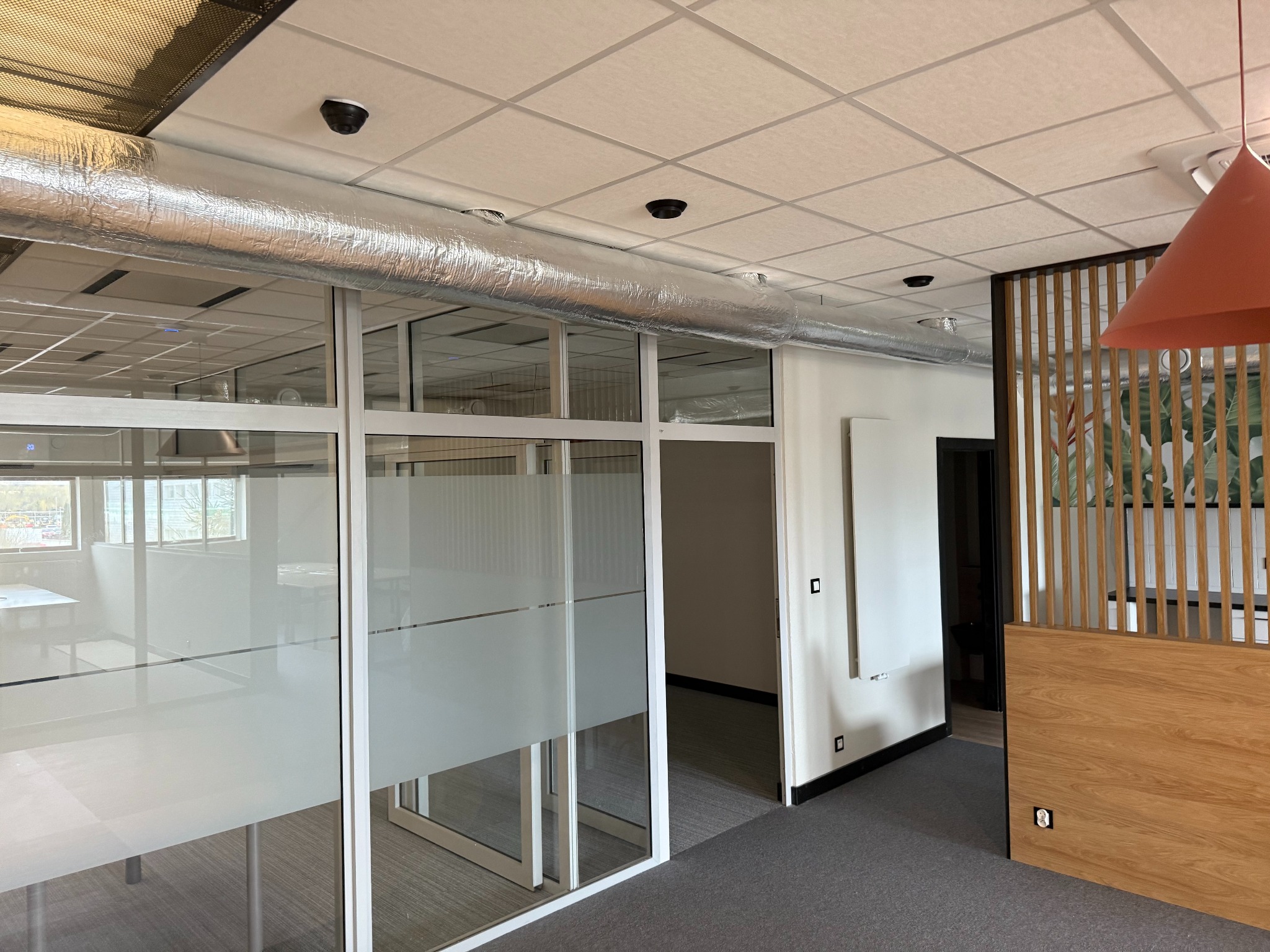 Biuro z widoczną instalacją wentylacyjną pod sufitem, szklane ścianki działowe z matową folią, widoczne czujniki dymu i oświetlenie.
