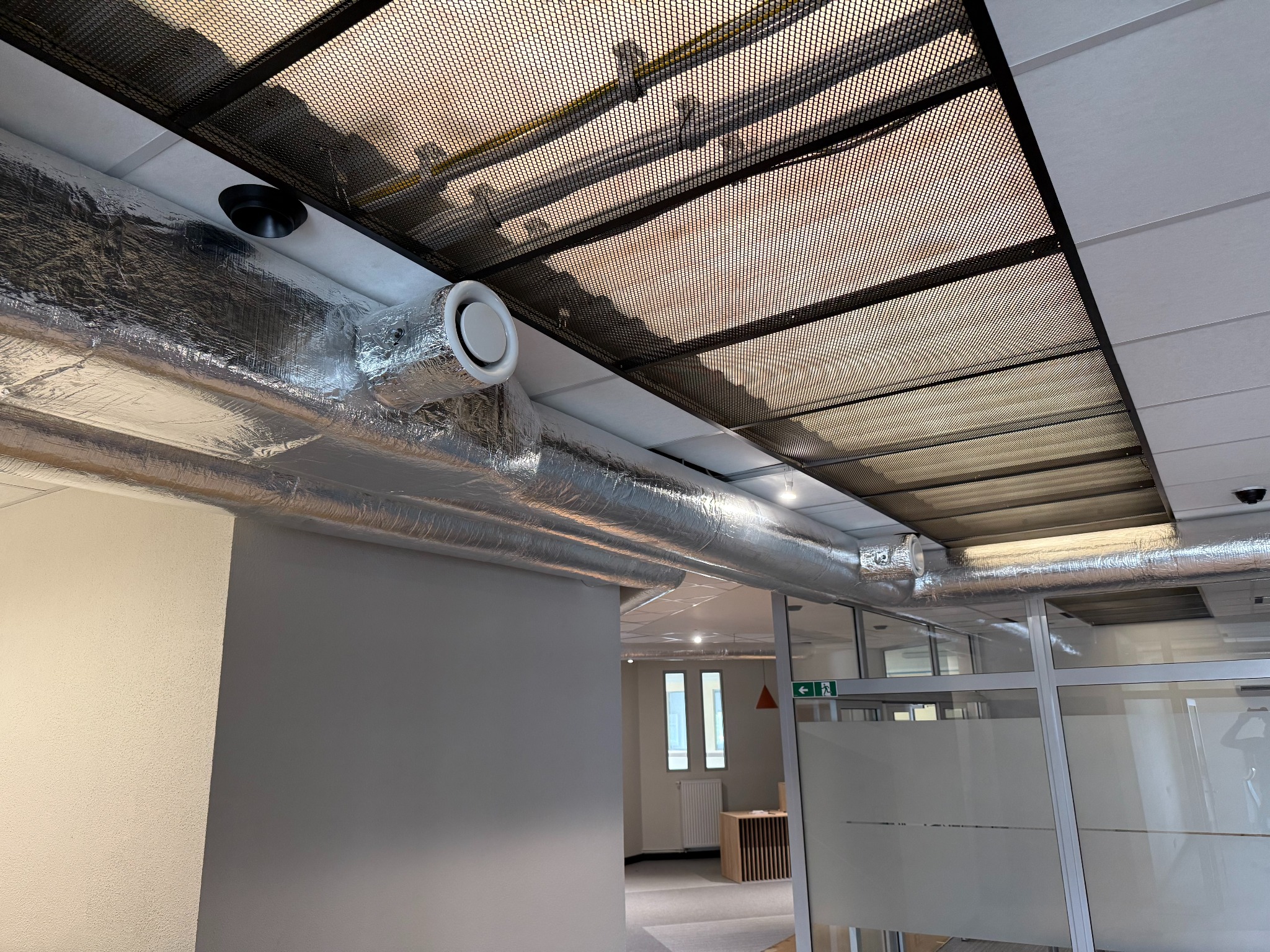 Instalacja wentylacyjna w biurze z widocznymi rurami w srebrnej izolacji i kratką maskującą pod sufitem. Widok na przeszklone biuro.