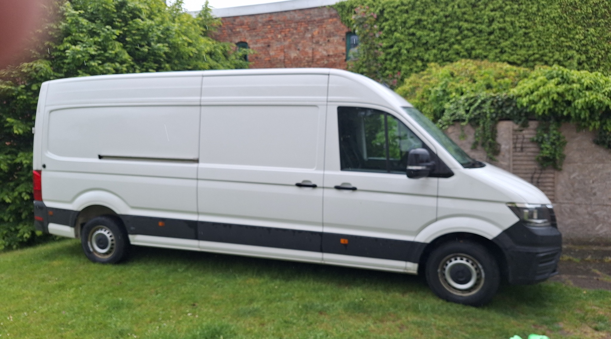 Biały, wysoki bus dostawczy marki Volkswagen Crafter zaparkowany na trawie przed budynkiem obrośniętym bluszczem. Widok z boku, lekko z góry.