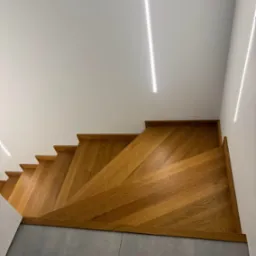 Kątowe ujęcie na dębowe schody ze stopniami w jodełkę, zintegrowane z szarą podłogą. Minimalistyczne oświetlenie LED podkreśla nowoczesny design.