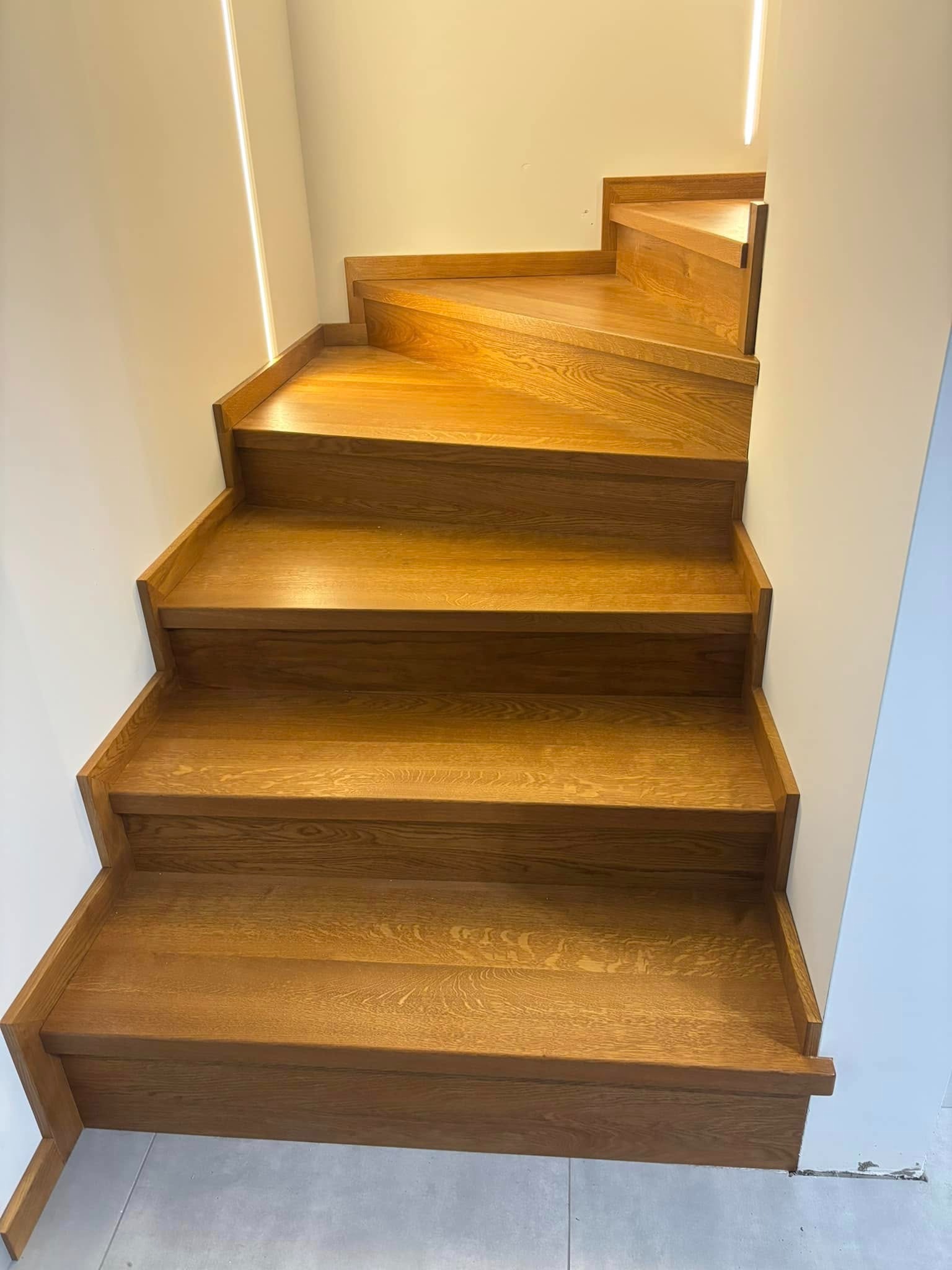 Kątowe ujęcie na jasne, dębowe schody z podświetleniem LED na ścianie. Stopnie z wyraźnym rysunkiem drewna, minimalistyczny design.