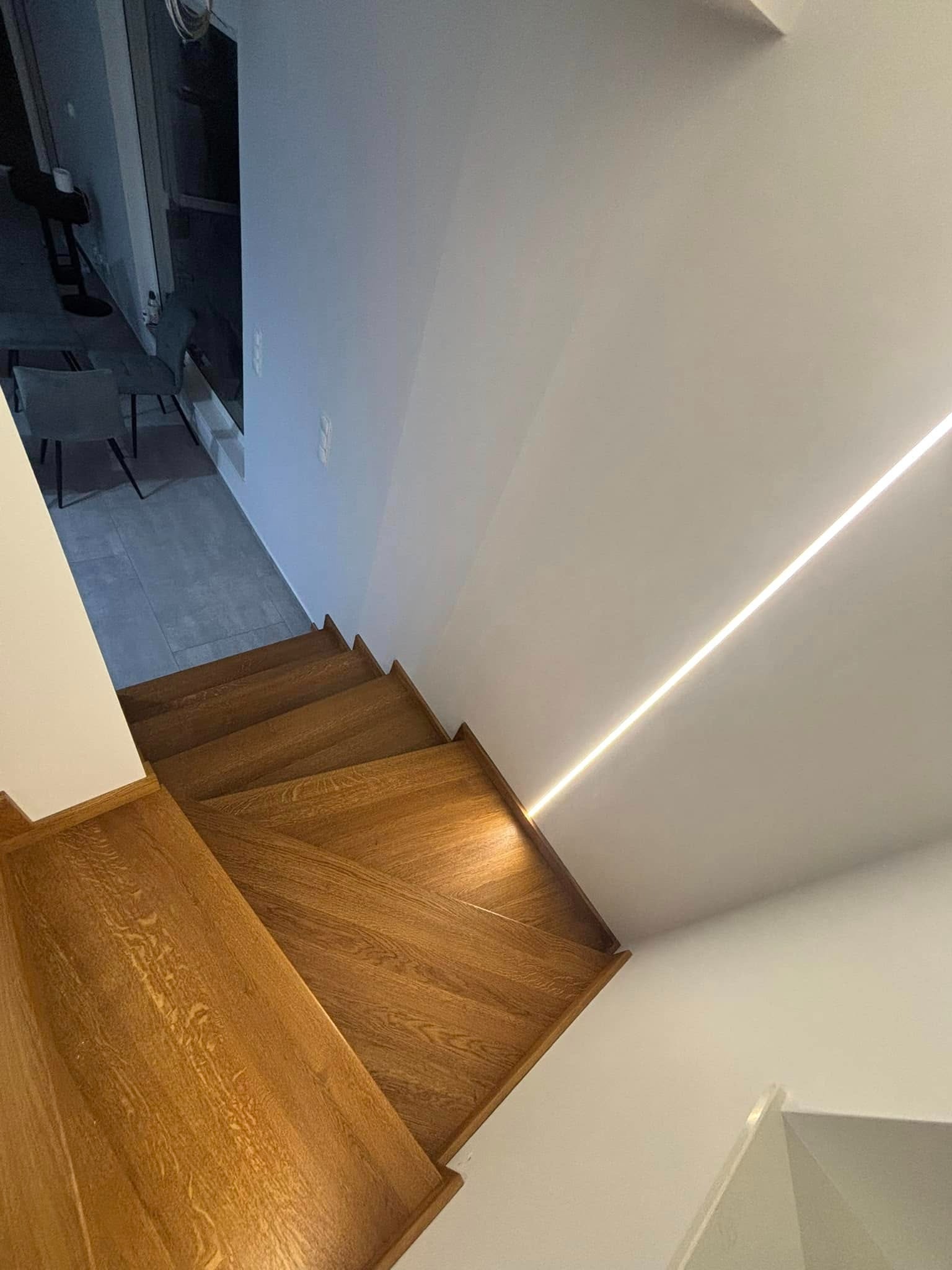 Nowoczesne schody z drewna dębowego, oświetlone listwą LED wzdłuż ściany. Widok z góry ukazuje strukturę drewna i minimalistyczny design.