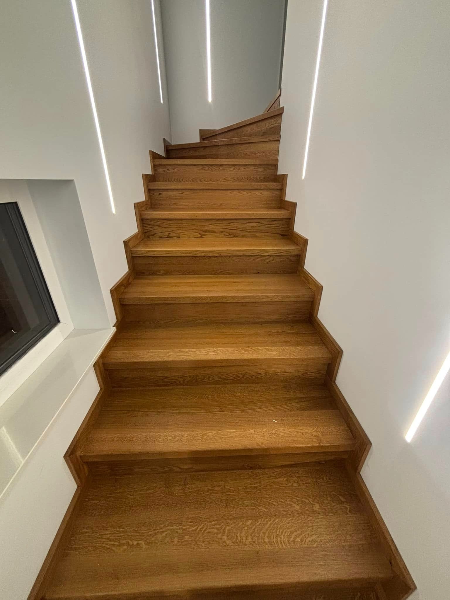 Nowoczesne schody z drewna dębowego z subtelnym oświetleniem LED wzdłuż ściany, widok z dołu, podkreślający minimalistyczny design i naturalną teksturę drewna.