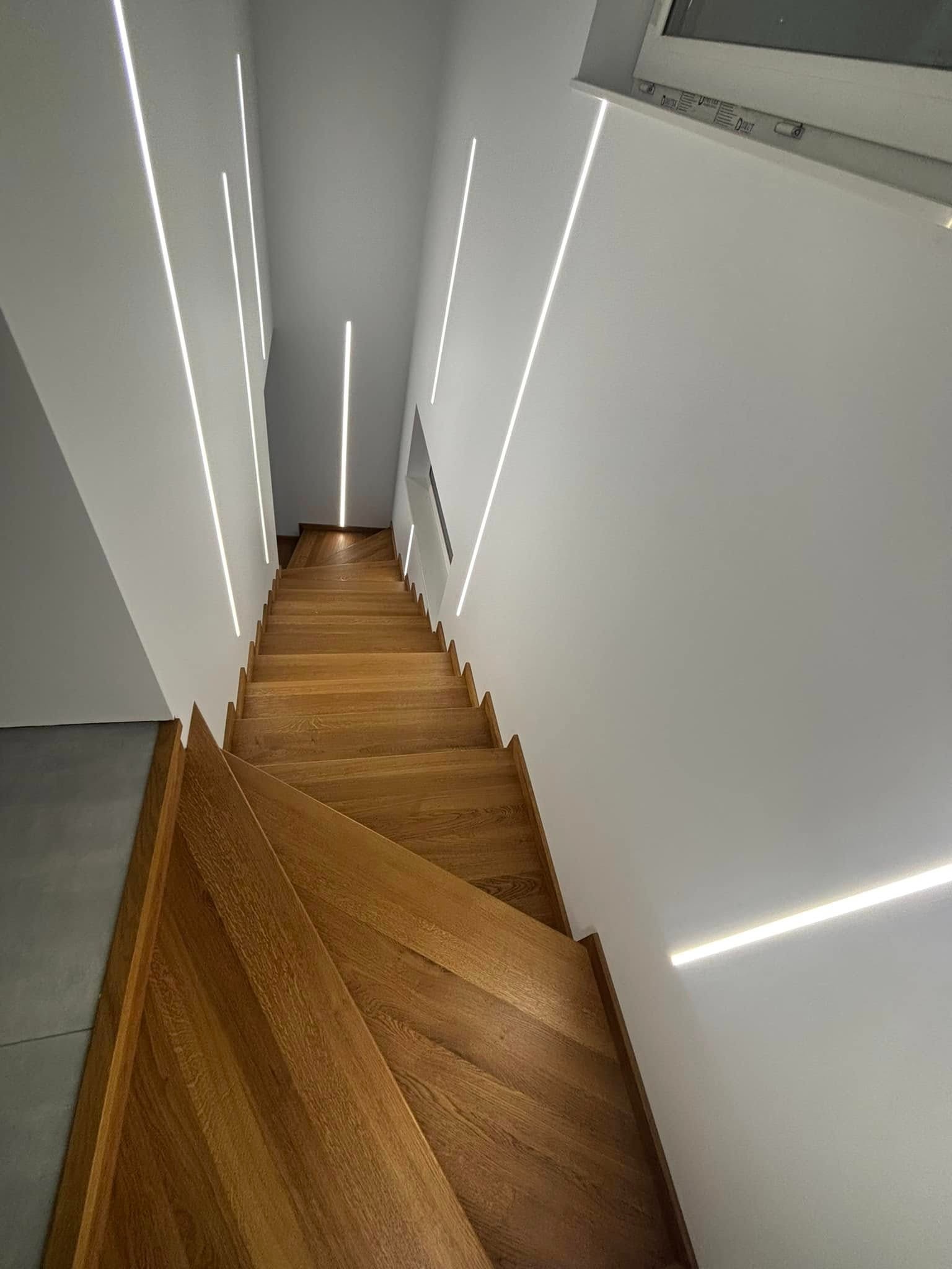 Nowoczesne schody z drewna dębowego, oświetlone subtelnymi listwami LED wzdłuż ścian. Minimalistyczny design i ciepłe światło tworzą elegancką przestrzeń.