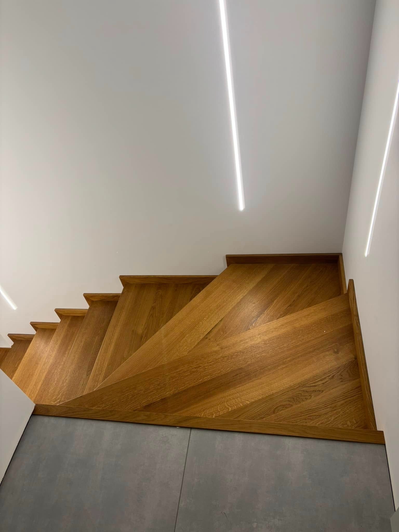 Kątowe ujęcie na dębowe schody ze stopniami w jodełkę, zintegrowane z szarą podłogą. Minimalistyczne oświetlenie LED podkreśla nowoczesny design.