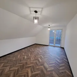 Poddasze z parkietem w jodełkę, białe ściany i czarne listwy przypodłogowe. Dwa nowoczesne, czarne plafony. Okno balkonowe z widokiem na zieleń.