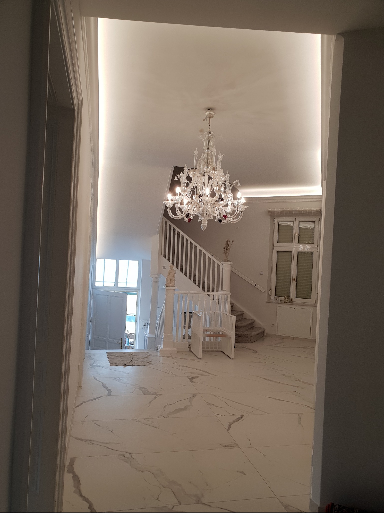 Jasne wnętrze domu z marmurową podłogą i schodami, oświetlone kryształowym żyrandolem, widziane z perspektywy korytarza. Białe ściany i elementy dekoracyjne.