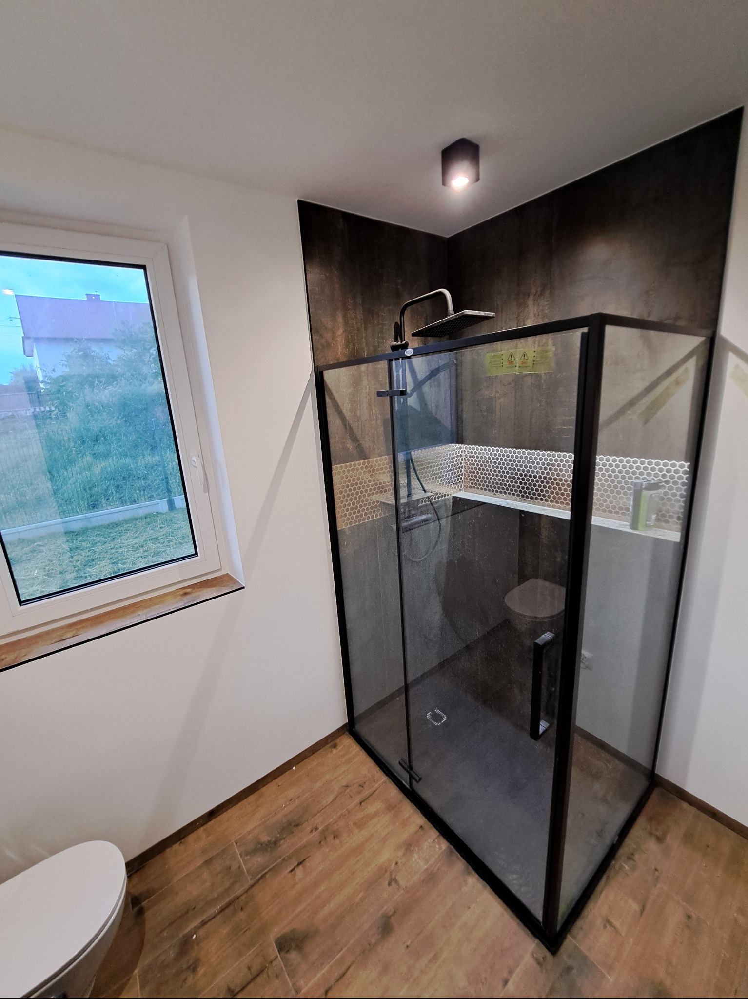 Nowoczesna kabina prysznicowa typu walk-in z czarnymi profilami, wykończona mozaiką. Widok z boku na drewnopodobną podłogę i fragment toalety. Okno z widokiem na zewnątrz.